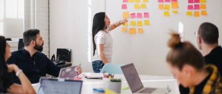 an asian woman points at some pink, yellow, and orange sticky notes on a whiteboard in a meeting with other people